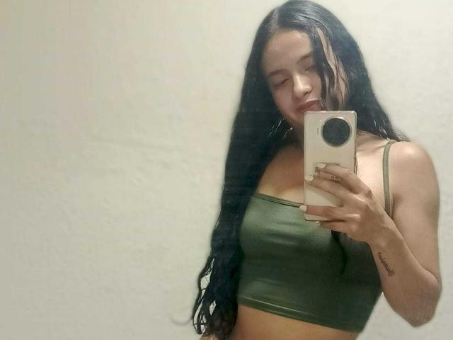SiennaTaylor, nua, com seios grandes e pele bronzeada, chupa um grande dildo vermelho enquanto está deitada em uma cama, vestindo meias pretas até a coxa e um colar de ouro, com um dildo marrom..