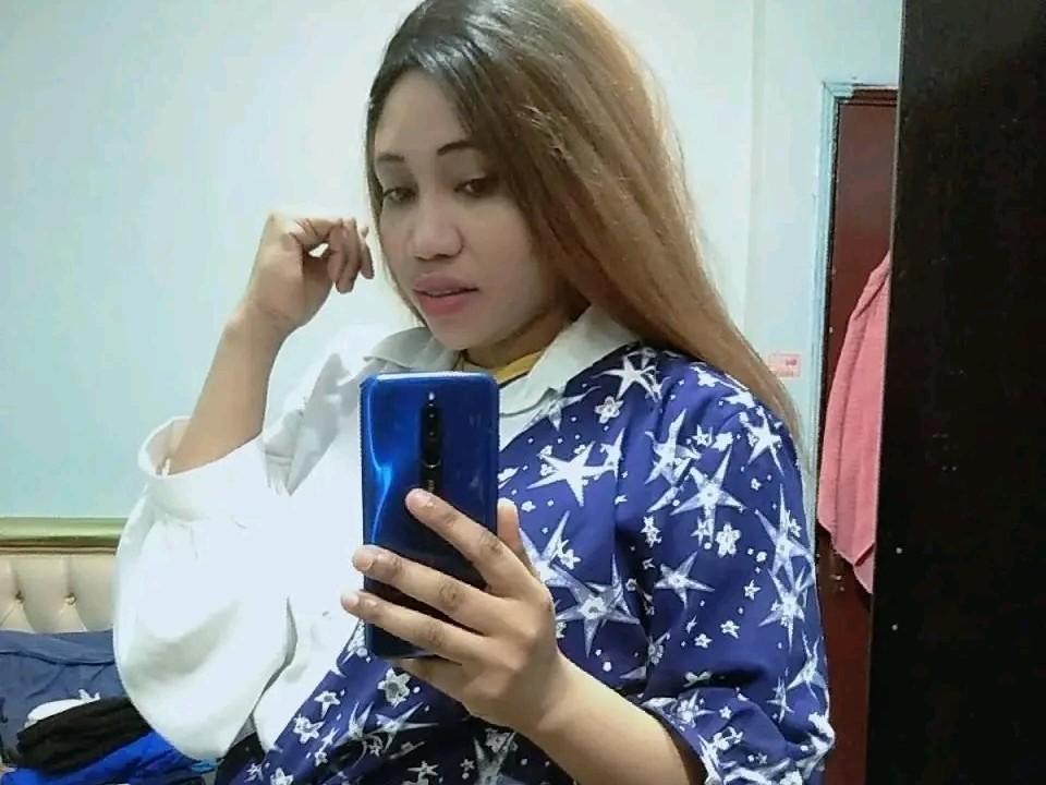 CrisyMarie, met lang zwart haar, draagt een zwart badpak met uitsparingen, waardoor haar billen en vagina zichtbaar zijn. Ze zit op een bed met zwarte kussens. Ze kijkt naar de camera..