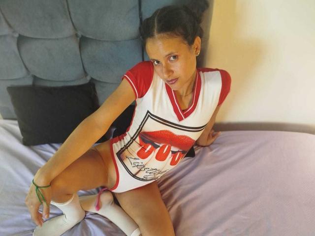 Dilay with dark skin, large boobs, and a curvy ass, sits on a black couch, wearing a white tied-up shirt, black shorts, white socks, and black shoes.