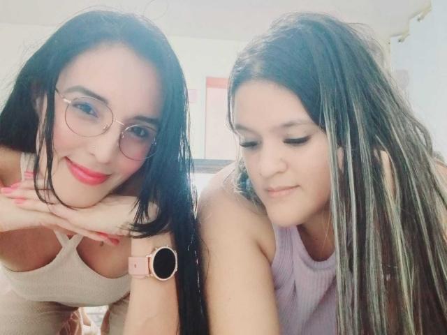 JenniferAndMia with dark hair, glasses, and pink top, holding a curvy JenniferAndMia with brown hair, purple top, and black jeans, standing under a ceiling fan.