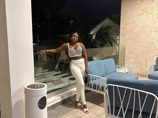 LeleFw stands smiling, dark-skinned, with sparkly bra, white pants, and white shoes, pointing outside, holding phone, blue cushions, and white patio furniture in the background.