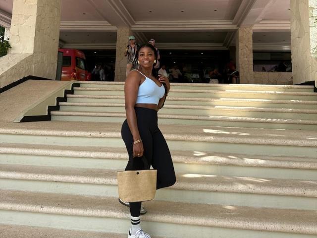 LeleFw with dark skin, small tits, wearing a blue crop top, black high-waisted pants, holding a tan bag, standing on wide stone steps, smiling, wearing white sneakers.