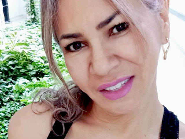 BrigiteWalsh has light brown skin, purple lipstick, and a cute smile with a slight frown. She has curly brown hair and is wearing a black top. Her tits are small.