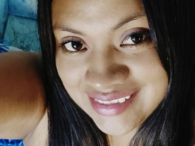 DahianaColeman's face with dark straight hair, brown eyes, pinkish-brown eyeshadow, and pink lipstick, smiling softly at the camera, showing her white teeth. Her skin is a warm brown.
