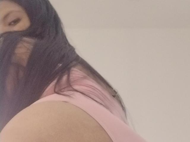 DahianaColeman with long black hair, pink underwear, and a visible butt. She's looking down at the camera with a subtle smile. Her face is partially hidden.
