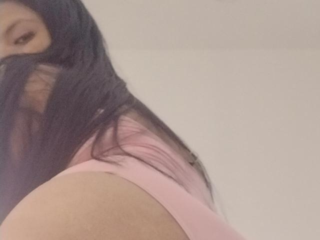 As nádegas de Dahiana Coleman, vestindo roupa íntima rosa, com seu longo cabelo preto cobrindo parte do rosto, olhando para baixo com um leve sorriso. A parte superior do seu corpo está parcialmente visível..