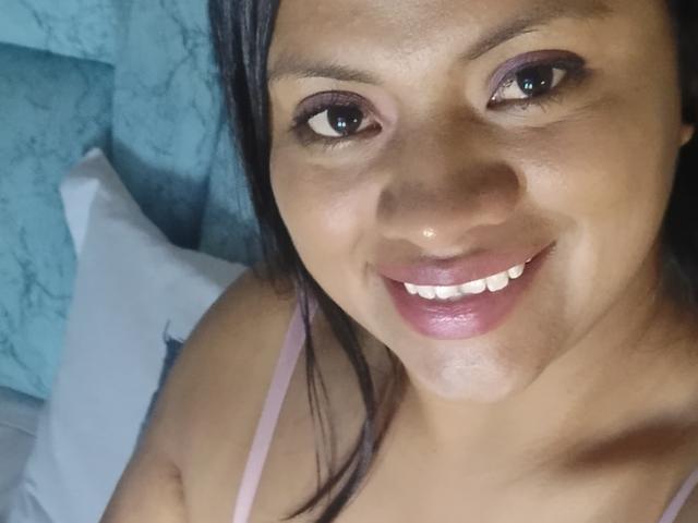DahianaColeman has dark brown skin, purple eyeshadow, and a pink smile with shiny lips, wearing a pink strap, looking up at the camera with a slight smile.