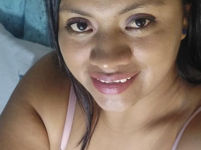Il viso di Dahiana Coleman, con la sua pelle colorata, occhi scuri con trucco, rossetto rosa e un leggero sorriso, indossa una canottiera rosa, con i capelli neri in primo piano..