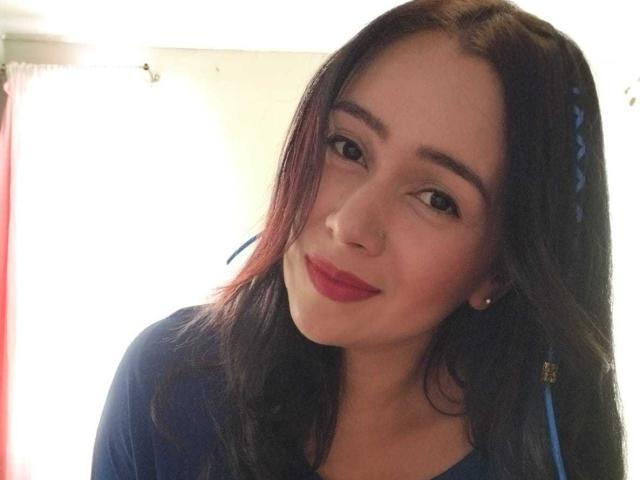 SarahShinny with dark brown hair, red lipstick, and blue earrings, smiling slightly, wearing a blue top, in a bright room. Her nose is pierced on the right side.