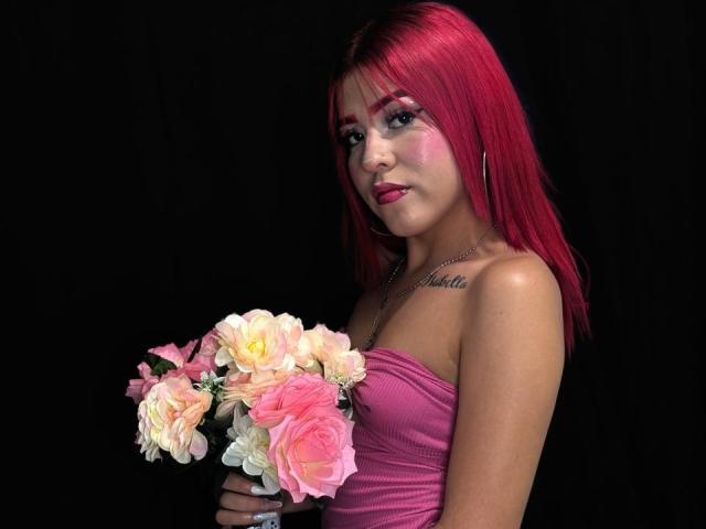ValeriGarcia with bright pink hair, wearing a strapless pink dress, sitting with legs crossed, showing her tits, a small tattoo above her right boob, and a necklace.