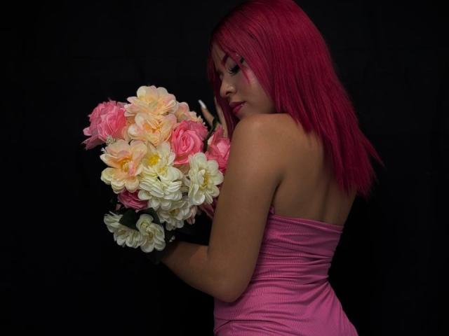 ValeriGarcia with pink hair, pink bra and panties, small tits, sitting on black bed, legs crossed, blue pillow behind, tattoo on chest, dark background, sexy, barefoot.