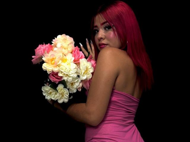 ValeriGarcia with pink hair, wearing a strapless pink dress, holding a bouquet of pink, white, and yellow flowers, looking over her shoulder, black background.