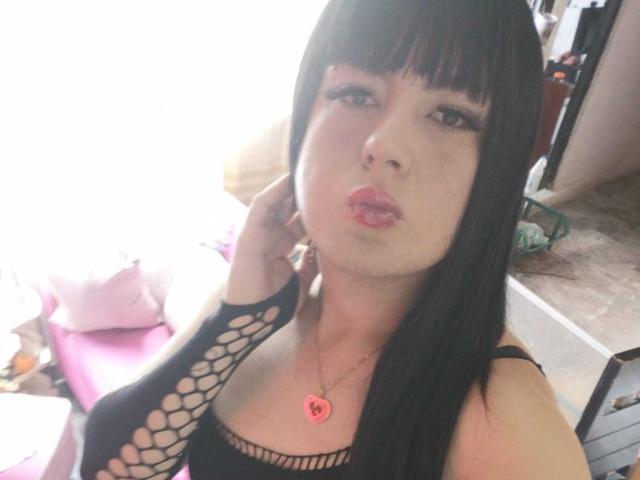 SashaPolet mit langen, blonden Haaren, trägt einen schwarzen Bikini-Oberteil, der ihre Brüste zeigt, und einen schwarzen String mit Spitzen-Hosenträgern. Sie sitzt auf einem Bett und hält ein Telefon..