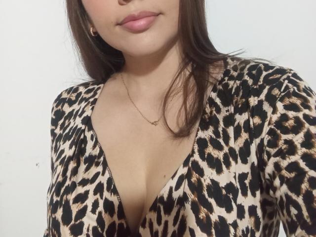 Ela Williams, com seios médios, veste um vestido de estampa de leopardo, usa um colar de ouro e um batom rosa discreto, e seu cabelo castanho cai sobre os ombros..