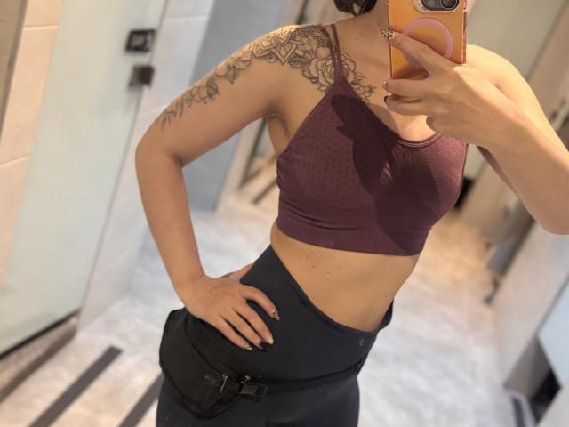 LaurentColin with tattoos on the shoulder, wearing a purple sports bra and black high-waisted pants, taking a mirror selfie, hand on hip, phone in orange case.