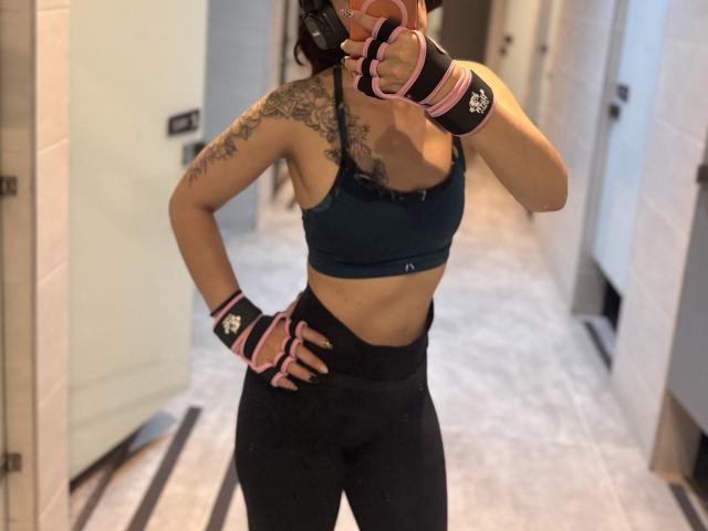 LaurentColin with tattoos on the right shoulder, wearing a teal sports bra, black high-waisted leggings, pink and black fingerless gloves, taking a mirror selfie in a locker room.