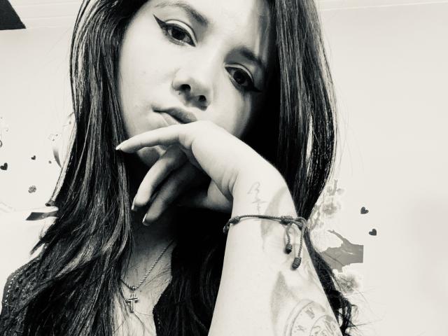 Black-and-white photo of Kkatalina with long dark hair, winged eyeliner, hand resting on chin, tattoo on wrist, necklace, and bracelet, looking slightly down with a pouty expression.
