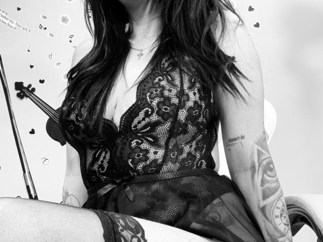 Zwart-wit foto van Kkatalina met lang donker haar, die een kant body suit draagt, waarbij haar borsten en tatoeages op haar arm zichtbaar zijn. Ze zit met een gitaar op de achtergrond..