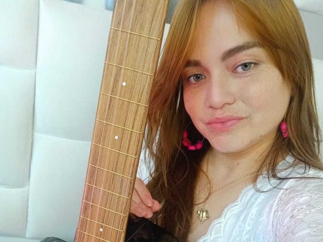 MoonRay69 houdt een basgitaar vast, met roze oorbellen, lichtbruin haar en een lichte frons. Ze draagt een witte kanten top en heeft een hartvormige ketting..