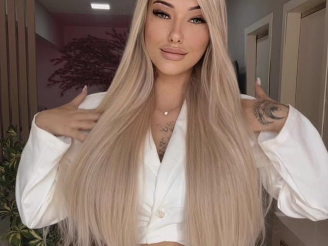 BarbPerversa has long blonde hair, a nose ring, and a tattoo on her right shoulder. She's wearing a white shirt, making a 'rock on' gesture with both hands.