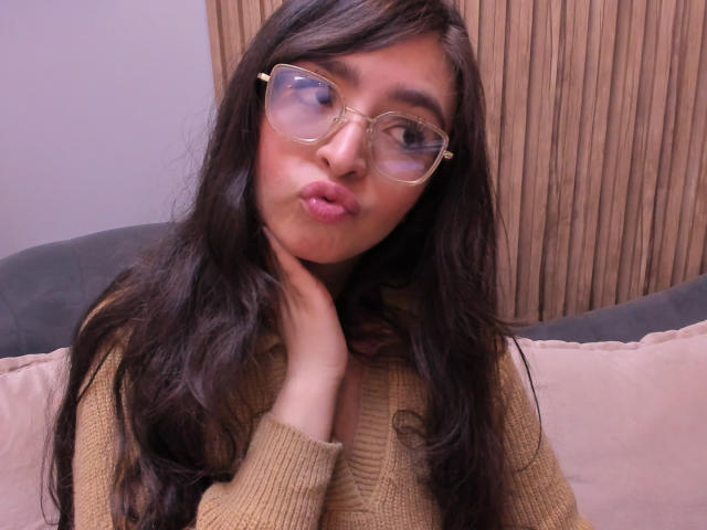 EvaMcAllister with long dark hair, pouty pink lips, and clear glasses, wearing a beige sweater, hand touching her neck, looking slightly to the side, in a casual selfie.