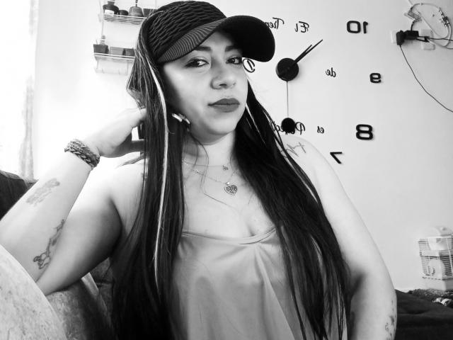 EvaLunaLatina with long dark hair, wearing a cap, tank top, and bracelets, looking at camera, showing tattoos on arm, sitting in black-and-white photo.