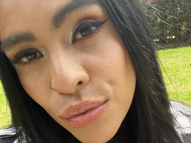 StellaBeckker's face, showing her pouty, glossy pink lips, dark eyeliner, and a nose ring, with her black hair and brown eyes. Her skin is light brown.