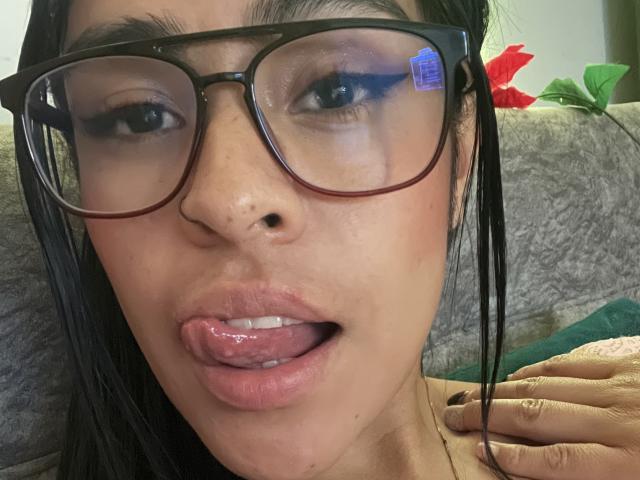 StellaBeckker's face with large black-framed glasses, dark hair, and brown skin. She's sticking out her wet tongue, showing her teeth, and has a small nose boogey.