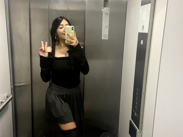 Elizaa with black hair, wearing a black off-shoulder top and black skirt, making a peace sign, standing in an elevator, holding a green phone. Visible tattoo on neck.