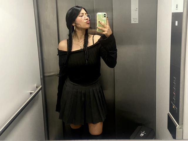 Elizaa with long black braids, off-shoulder black top, and black skirt, taking a mirror selfie in an elevator, pouting with her phone in hand, showing her tits.