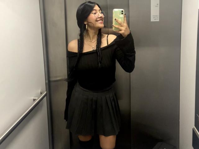 Elizaa with long black braids, smiling, taking a selfie in an elevator, wearing an off-shoulder black top and a pleated black skirt, holding a green phone.