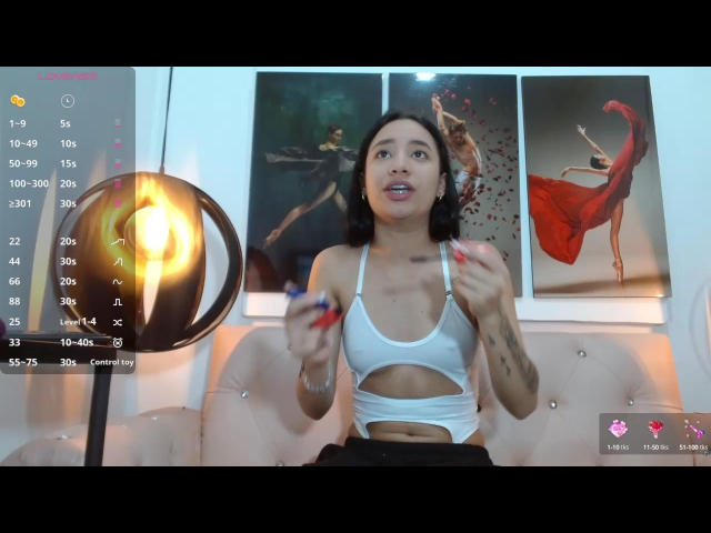 SelenaRamirez with medium tits, wearing a white cut-out top, holding red nails, sitting on a pink tufted couch, tattooed arms, looking up with mouth open, dark curly hair.