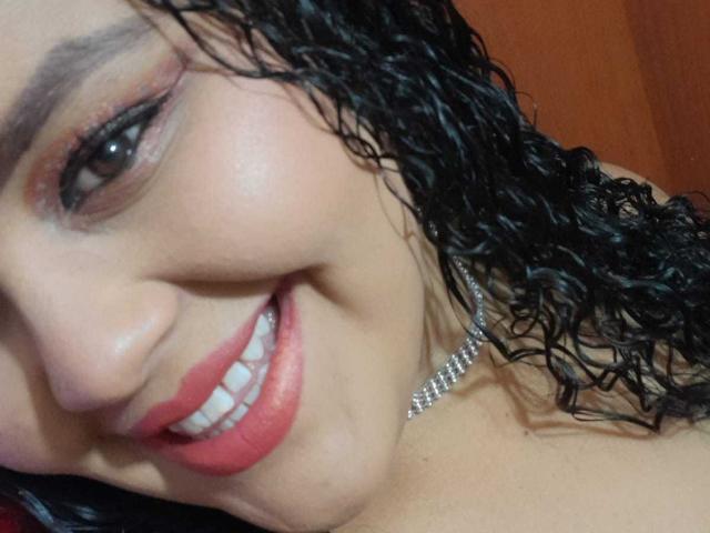 MelannieCarter's smiling face with red lipstick, curly black hair, and silver necklace. She's got a cute smile and brown eyes, showing her white teeth. Her tits are partially visible.