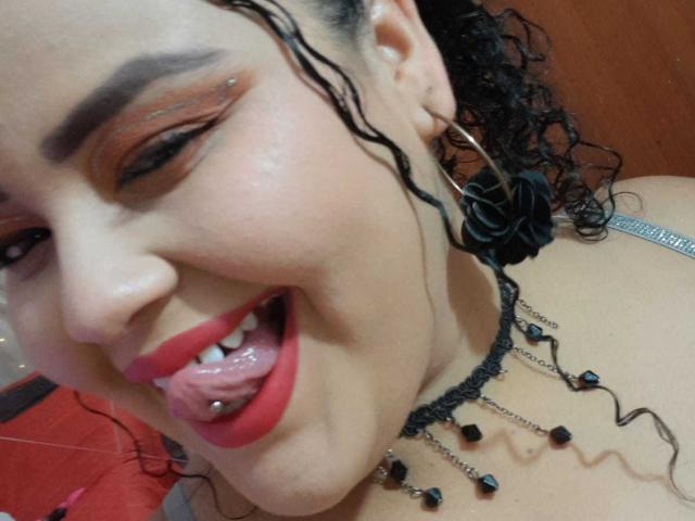 Le visage de Melannie Carter, la langue tirée avec un piercing noir, du rouge à lèvres, des cheveux noirs bouclés, des boucles d'oreilles et un collier avec des perles noires. Elle sourit et porte du maquillage..