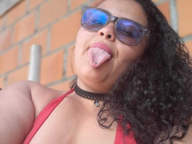 MelannieCarter with curly black hair, blue-tinted sunglasses, red top, black choker, and tongue out, showing a tongue piercing. Boobs visible, slightly above the top.