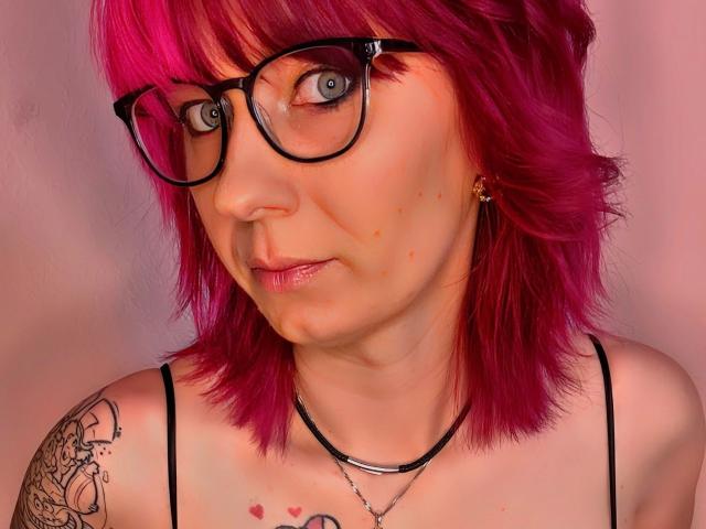 Ladyminniemaus-hot has pink hair, black glasses, and a tattoo on her right shoulder. She's wearing a black spaghetti strap top, looking at the camera with blue eyes.