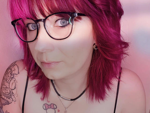 Ladyminniemaus-hot with pink hair, black glasses, and tattoos, showing a surprised expression, has pale skin, small tits, and a black choker necklace.