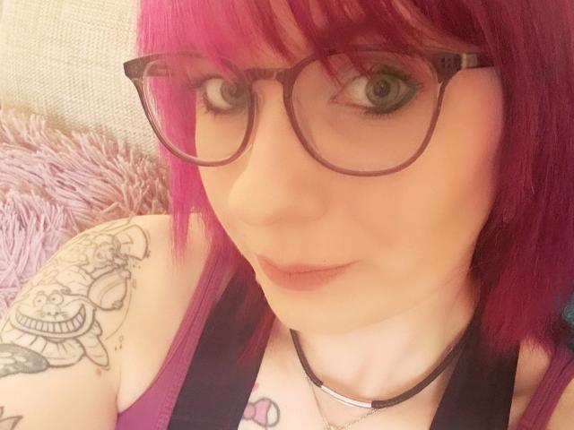 Ladyminniemaus-hot with pink hair, glasses, and tattoos, showing her face and upper chest, wearing a black top, necklace, and pink lipstick, with a slight smile.
