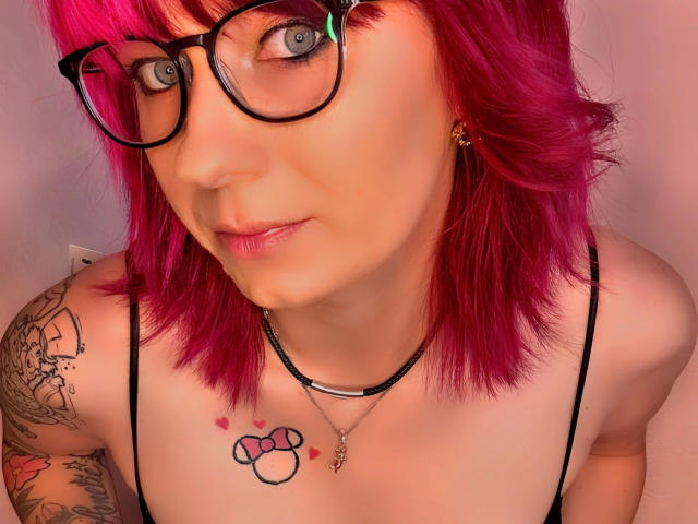 Ladyminniemaus-hot with pink hair, glasses, and tattoos, showing tits, wearing a black strap top, and a necklace, looking into the camera with a subtle smile.