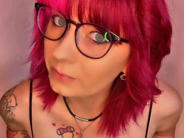 Ladyminniemaus-hot with bright pink hair, black glasses, blue-green eyes, and tattoos on her shoulder and chest. She's wearing a black strap top and a black necklace.