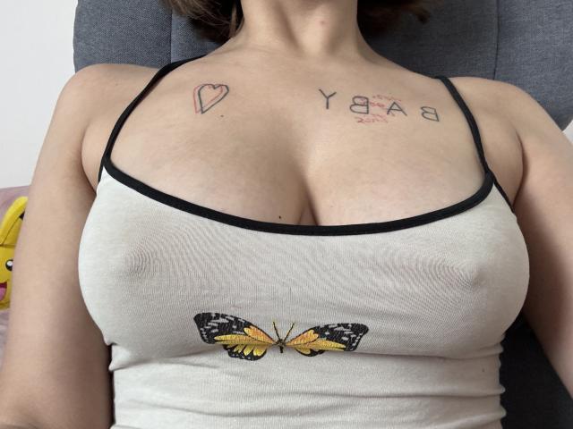 Les grandes seins de Kametia sont visibles sous un débardeur blanc avec des bretelles noires, orné d'un motif de papillon, et elle a des tatouages d'un cœur et de "YGA" sur sa poitrine..