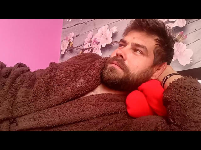 Mhatyas with a brown fluffy robe, red gloves, and a beard, lying down, looking to the side, short dark hair, pink and purple lighting, floral wall art in background.