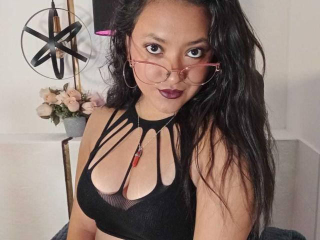 SafiMontiel with long black hair, wearing a black strappy top, red glasses, and dark lipstick, showing her tits and a red pendant. Background includes a pink flower vase.