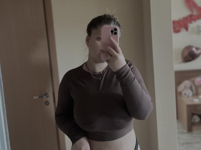 Madrinia with brown long-sleeve crop top, showing her belly, holding a pink phone, taking a mirror selfie, wearing black panties, light skin, brown hair, small earrings.