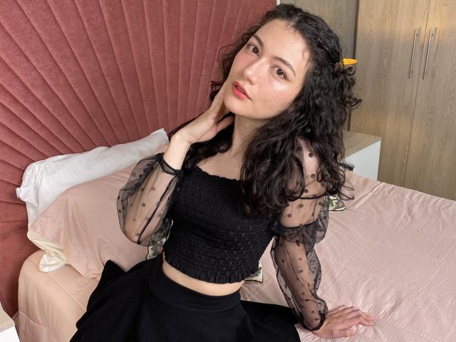 AmeliaTorres with curly black hair, wearing a sheer black polka dot top and black skirt, sitting on a pink bed, touching her chin, with a relaxed, slightly sultry expression.