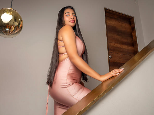 NikkiSpice, with long dark hair, wearing a tight pink dress, stands on a staircase, hands on the railing, showing her ass and back, looking over her shoulder.