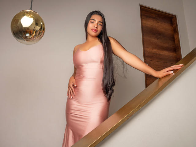 NikkiSpice stands on a staircase, wearing a tight pink dress, long black hair, hand on her ass, gold railing, looking down, smiling, under a reflective hanging light.
