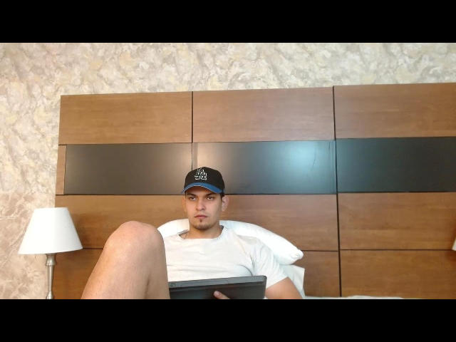 ErickStonee with a serious face, wearing a black cap, white t-shirt, and holding a tablet, sitting against a wooden headboard with a white lamp on the left.