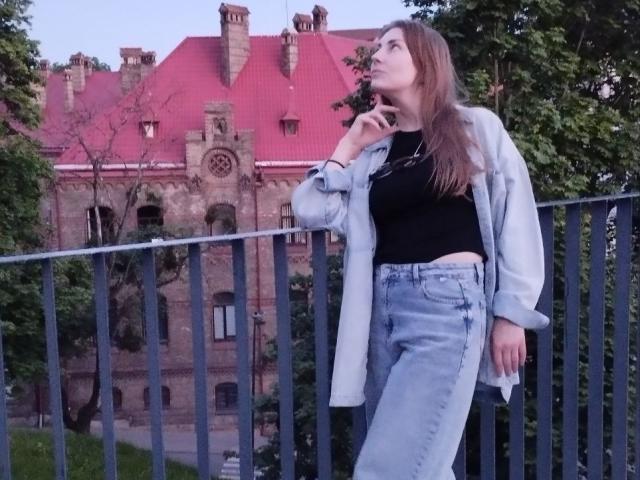 BlackMango with long brown hair, wearing a black top, blue jeans, and a light gray jacket, standing by a metal railing, looking up thoughtfully at a red-roofed building.