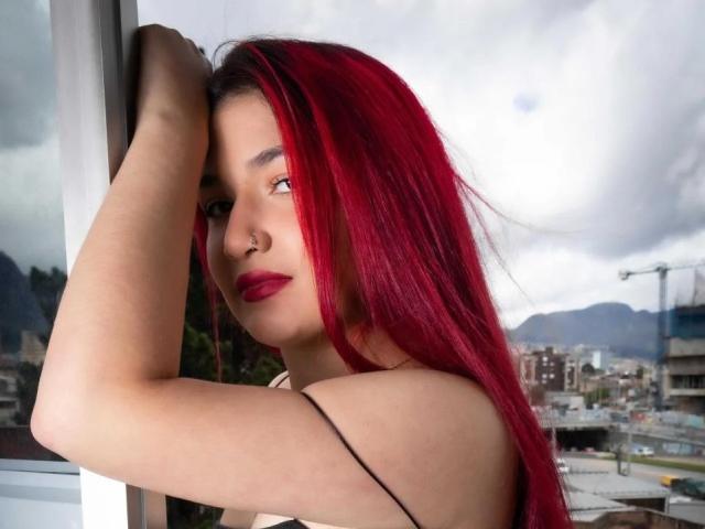 AmyRousse with bright red hair, nose piercing, and red lipstick, arms raised, looking over shoulder, showing her tits, standing by a window, cityscape in background.